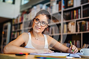 Portrait of happy young female student taking notes from a book at college library.