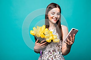 Portrait of a happy young girl in dress use mobile phone while holding big bouquet of yellow tulips isolated over color background