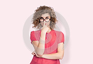 Portrait of a happy young girl in a red dress holding glasses on her nose with her finger, on light red background