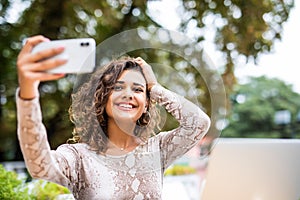 Portrait of a happy young girl taking selfie with mobile phone while sitting with laptop computer at a cafe outdoors