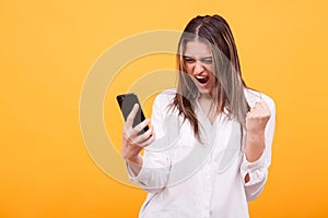 Portrait of a happy young girls holding mobile phone and celebrating isolated over yellow background