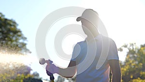 Portrait of happy young guy gardener holding in hand hose and watering green plant in garden. Joyful man spraying water