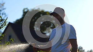 Portrait of happy young guy gardener holding in hand hose and watering green plant in garden. Joyful man spraying water