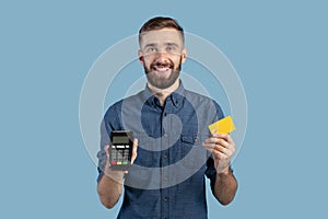 Portrait of happy young guy holding credit card and payment terminal, amiling and looking at camera over blue background