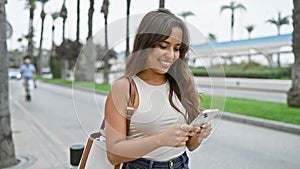 Portrait of a happy young hispanic woman, with a beautiful hairstyle, joyfully using her smartphone on an urban street, cheerfully