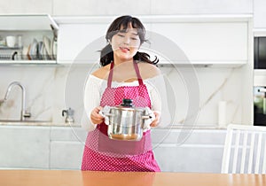 Portrait of happy young housewife in apron posing at kitchen, holding bowl of soup