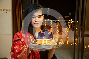 Portrait of Happy Young indian beautiful woman hold plate/thali with diya/clay oil lamps wearing traditional dress,standing indoor