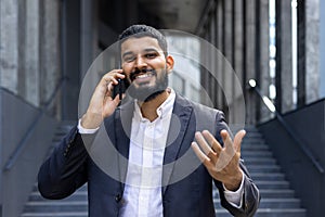 Portrait of a happy young Indian businessman talking on the phone on the street, gesturing with his hands and smiling at