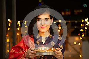 Portrait of Happy Young indian gorgeous woman hold plate/thali with diya/clay oil lamps wearing traditional dress,celebrates