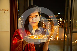 Portrait of Happy Young indian gorgeous woman hold plate/thali with diya/clay oil lamps wearing traditional dress,celebrating