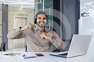 Portrait of a happy young Indian man sitting at a desk in the office, working on a laptop, looking smiling at the camera