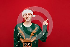 Portrait of happy young man in christmas sweater and santa hat showing thumbs up, looking into camera and smiling. The guy in