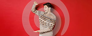 Portrait of happy young man in christmas sweater, showing large present, smiling and looking amazed at cool gift