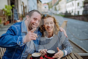 Portrait of happy young man with Down syndrome with his mother sitting at cafe outdoors and talking.