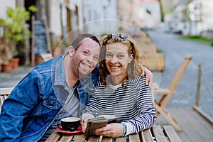 Portrait of happy young man with Down syndrome with his mother sitting at cafe outdoors and talking.