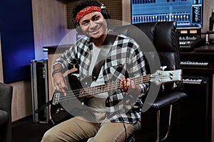 Portrait of happy young man, male artist in headphones smiling aside, playing guitar while sitting in recording studio