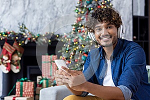 Portrait of happy young man with phone and headphones, sitting on sofa in living room near Christmas tree, smiling and