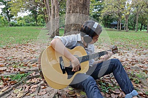 Portrait of happy young man playing acoustic guitar and leaning a tree at the outdoor park.