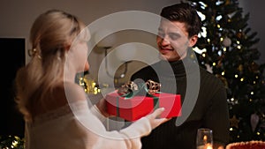 Portrait of happy young man receiving gift box from loving woman sitting at dinner table at home on Christmas Eve near