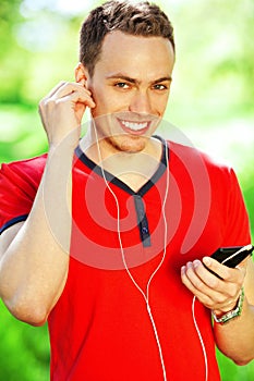 Portrait of happy young man in red t-shirt listening to music on smartphone in the park. Handsome muscular guy in casual clothing