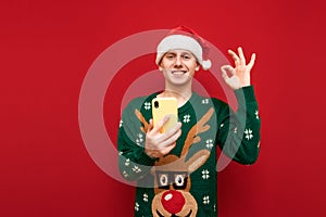 Portrait of happy young man in santa claus hat and christmas sweater stands on red background with smartphone in hand, looks into