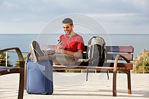 Happy young man sitting on bench with luggage and listening to music with mobile phone and earphones