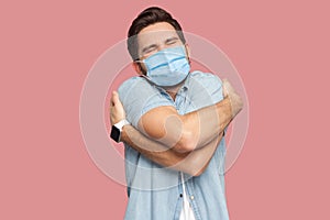 Portrait of happy young man with surgical medical mask in blue shirt standing and hugging himself with closed eyes and smile