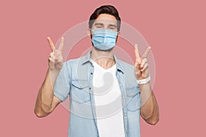 Portrait of happy young man with surgical medical mask in blue shirt standing with victory or peace hand sign gesture and looking