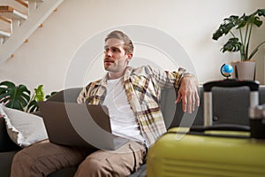 Portrait of happy young man, tourist booking his holiday with travel agency, looking at laptop, packed suitcase to go on