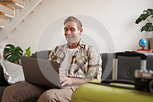 Portrait of happy young man, tourist booking his holiday with travel agency, looking at laptop, packed suitcase to go on