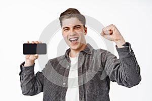 Portrait of happy young man winning on mobile phone, yelling from victory, fist pump and smiling pleased, showing