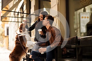 Portrait of happy young man and woman have walk outdoor with their pet, feed it with something delicious, sit on chairs