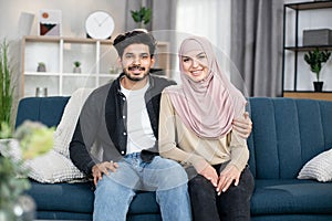 Portrait of happy young muslim couple sitting together at home on blue soft couch at cozy living room, smiling to camera