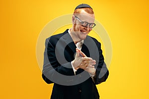 Portrait of happy young redhead Jewish man in glasses and yarmulke cheerfully posing against yellow studio background