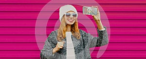 Portrait happy young smiling woman taking a selfie by smartphone wearing a knitted white hat on colorful pink background