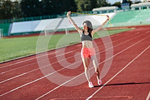 Portrait of happy young sports brunette woman in black top and rose shorts outdoors on stadium holding skipping rope over the head