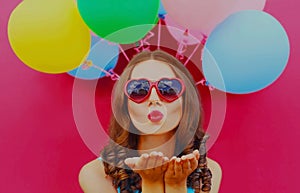 Portrait happy young woman blowing her red lips sending air kiss with colorful balloons on a pink background