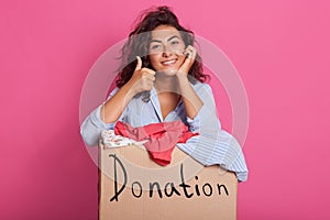 Portrait of happy young woman with clothes donation standing over rosy background, wearing casual outfit, keeps one hand under