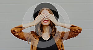 happy young woman covering her eyes with her hands wearing a black round hat over gray background