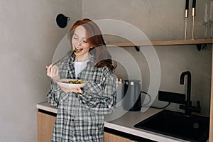 Portrait of happy young woman eating salad with organic vegetables, enjoying healthy diet, standing in modern kitchen