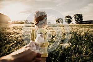 Portrait of a happy young woman holding hand of her boyfriend while walking by a wheat field at sunset