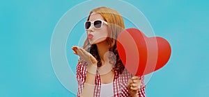 Portrait of happy young woman holding red heart shaped balloon and blowing lips sending sweet air kiss on blue background