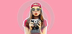 Portrait of happy young woman photographer taking picture on film camera and blowing her lips wearing baseball cap on pink