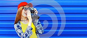 Portrait of happy young woman photographer taking picture on film camera wearing red baseball cap on blue background, banner blank