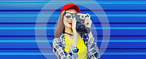 Portrait of happy young woman photographer taking picture on film camera wearing red baseball cap on blue background, banner blank