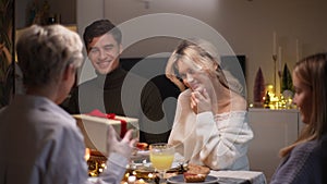 Portrait of happy young woman receiving gift from loving mother-in-law sitting with family at dinner feast table at home