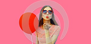 Portrait of happy young woman with red balloon blowing her lips wearing heart shaped sunglasses on pink studio background