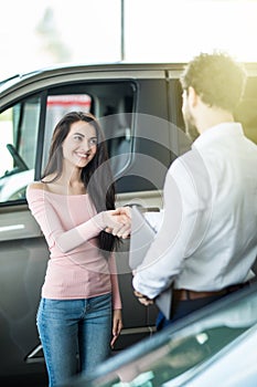 Portrait of happy young woman shaking hands with car salesman after buying new luxury car in dealership showroom