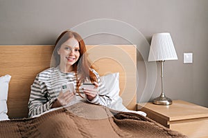 Portrait of happy young woman shopping online with credit card and smartphone sitting in bed at home, looking at camera.