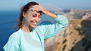 Portrait of happy young woman standing on cliff edge against amazing seascape and sky, hair blowing in the wind